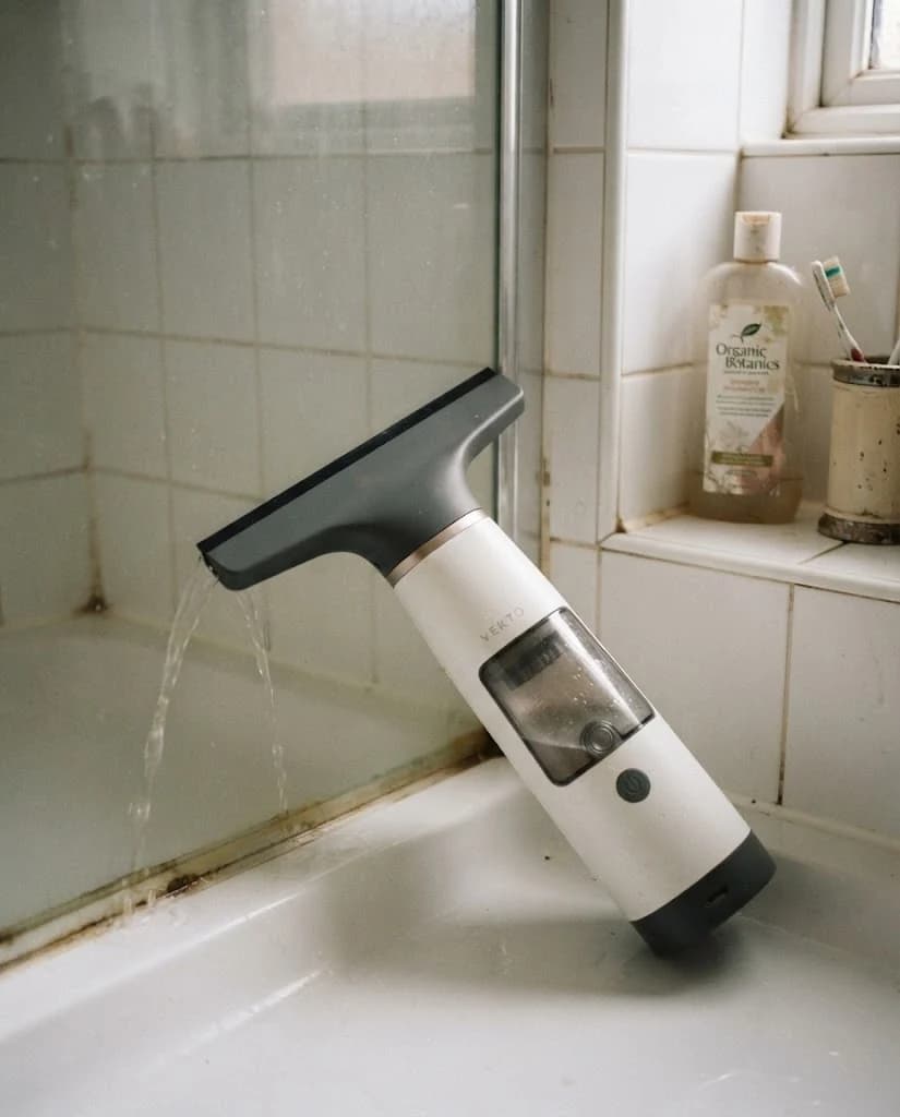 Vekto Window Vac W10 charging by USB-C on a bathroom windowsill beside a phone charger